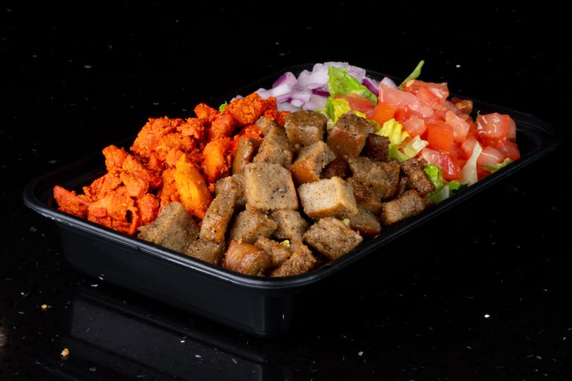 Takeout container with red chicken, brown meat, red onions, lettuce, and diced tomatoes on a black surface.